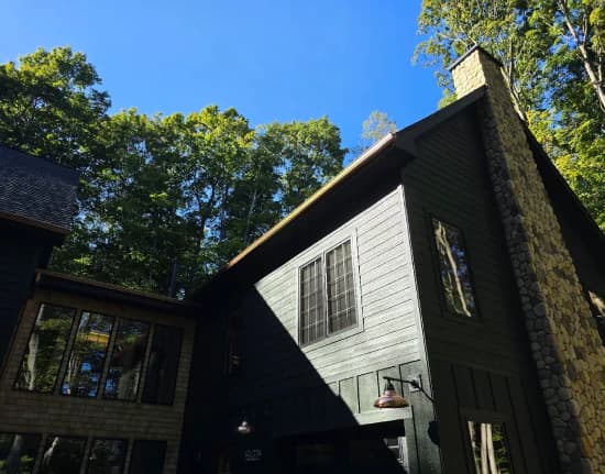 Tall dark-sided home with stone chimney and new gutters under a blue sky